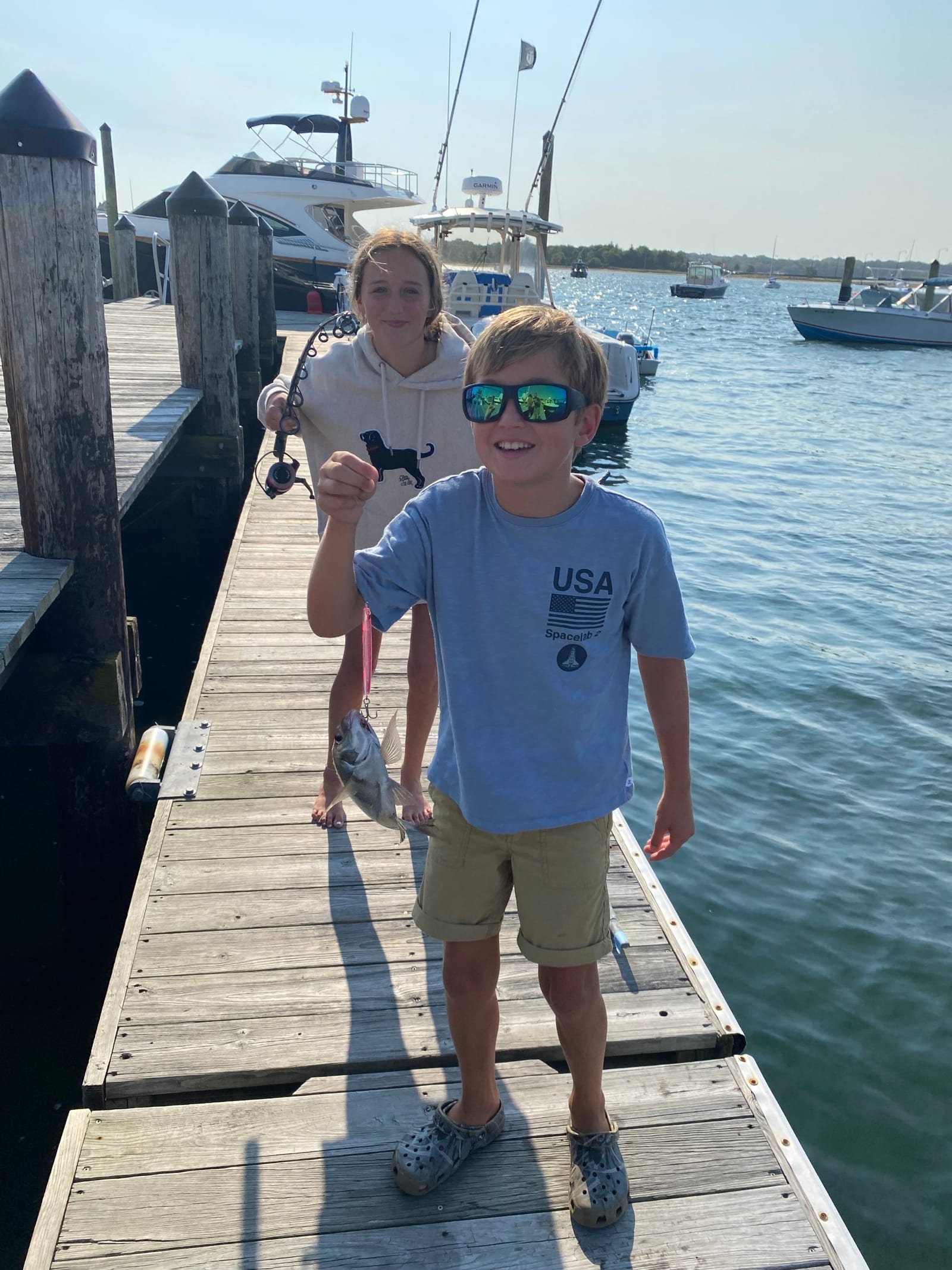 A boy on a Padanaram dock holding up a scup on a hook, his sister with the rod behind him, boats on the harbor.